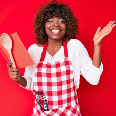 Young african american woman wearing professional baker apron reading cooking recipe book celebrating victory with happy smile and winner expression with raised hands